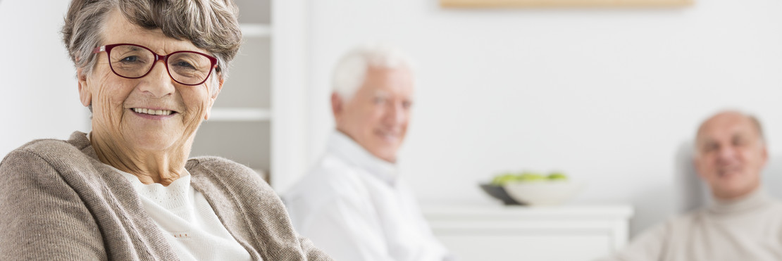 eine alte Frau lächelt in die Kamera. Im Hintergrund schauen zwei lächelnde alte Männer.
