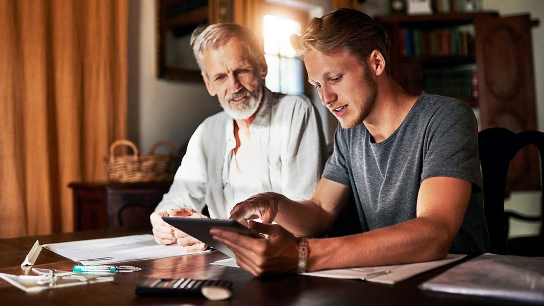 Ein Vater und sein erwachsener Sohn sitzen an einem Schreibtisch mit Tablet, Unterlagen und Taschenrechner. 