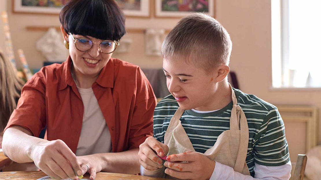 Ein Kind mit Downsyndrom malt gemeinsam mit einer erwachsenen Frau an einem Bild. 