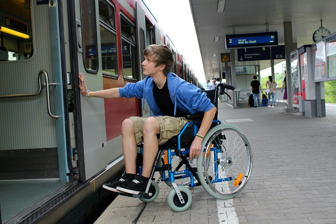 Junge im Rollstuhl auf einem Bahnsteig vor einem Zug. 