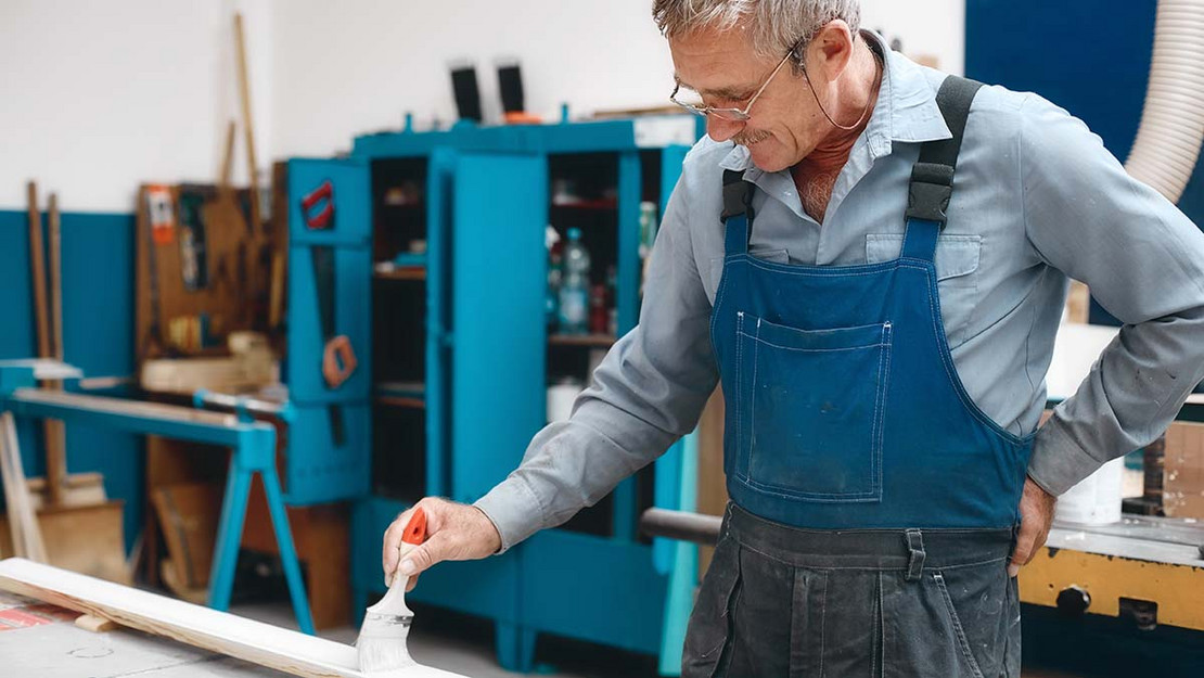 Ein älterer Handwerker streicht eine Holzlatte mit einem Pinsel an. 